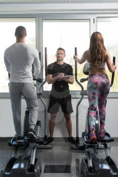 Group Of People Exercising On Elliptical Walker In Gym Or Fitness Club While Personal Trainer With Clipboard Watching Them - Group Of Woman And Men Exercising To Gain More Fitness