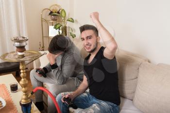 Two Young Gamers Sitting Together On Sofa And Playing Video Games At Home