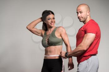 Muscular Man Measuring Partner Waist With Tape Measure - Fitness Couple Diet On Grey Background