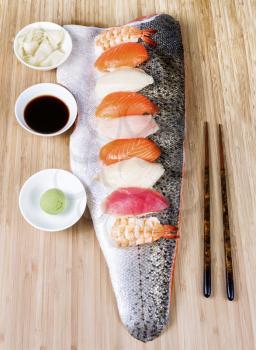 Vertical display of several pieces of assorted sushi placed on large salmon fillet with soy sauce, spicy Japanese mustard, ginger, and chop sticks on wooden bamboo board
