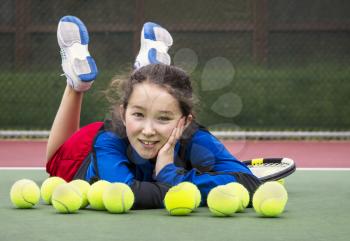 Horizontal portrait of smiling teenage girl tennis player laying on the court behind a row of tennis balls with head resting in her hand and feet in air