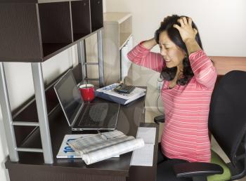 Mature Asian woman express frustration while doing income taxes with tax form booklet, calculator, coffee cup, glasses and computer on desk