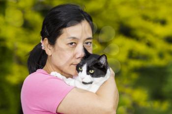 Horizontal photo of a mature woman holding a black and white cat while looking forward with blurred out green and yellow trees in background