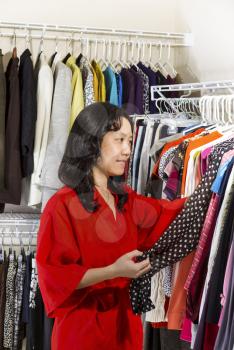 Vertical portrait of mature Asian woman, dressed in red bath robe, in walk-in closet inspecting her clothing before wearing