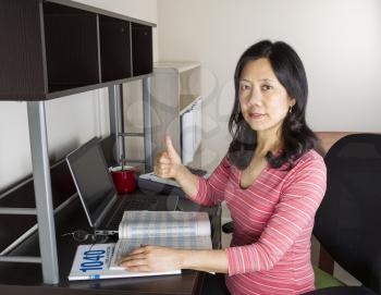 Mature Asian woman giving thumbs up with income tax tables booklet, computer, coffee cup and glasses on desk