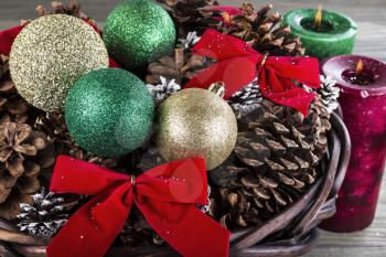 Holiday basket containing red ribbons, dry pine cones, gold and green ornaments and candles with flame in background