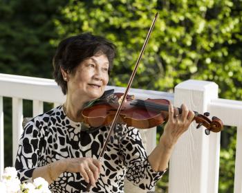 Horizontal photo of Senior Asian woman sitting down while playing the violin outdoors with green trees in background
