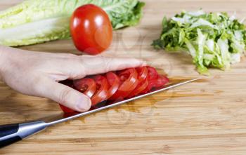 Horizontal photo of female hand with sliced tomato on knife blade, lettuce on natural bamboo cutting board