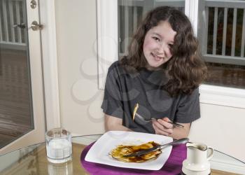 Young adolescent girl with piece of pancake on fork with big smile on her face and single large pancake on white plate with maple syrup on top