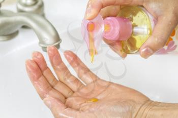 Horizontal photo of female hands putting liquid wash soap, from bottle, into hand with bathroom sink and faucet in background