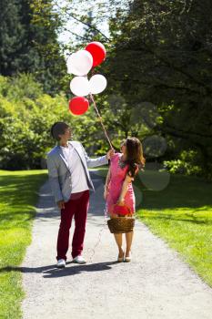 Vertical photo of young adult couple dressed in formal attire looking at several balloons while holding picnic basket with walking path, green grass and trees behind them  