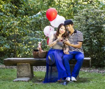 Photo of young adult couple sitting on log bench, looking forward, with glasses filled with red wine being held in their hands with balloons, green grass and trees in background 