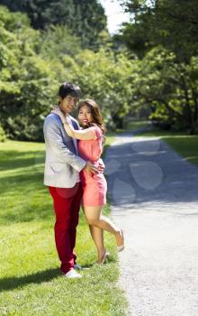 Vertical photo of young adult couple dressed in formal attire holding each other with walking path, green grass and trees behind them  