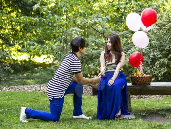Horizontal photo of young adult man, on one knee with single red rose, proposing to his lady with balloons, basket of fruit and trees in background 