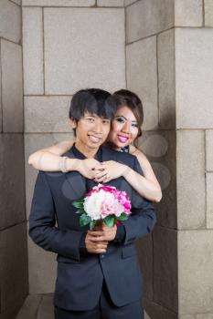 Vertical photo of young adult couple, man holding flowers, with lady holding him on stone wall in background 