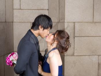 Closeup photo of young adult couple kissing with hidden flowers behind the man on a stone wall in background 