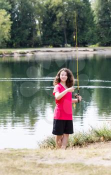 Vertical photo of young girl, looking forward, holding small fish that she caught with lake and trees in background 