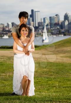 Vertical photo of young adult couple, facing forward, holding each with city of Seattle and harbor in Background