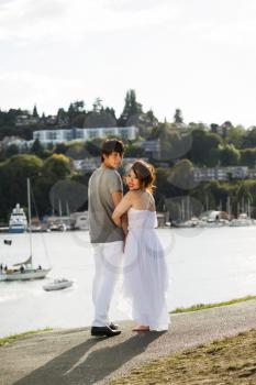 Vertical photo of young adult couple, looking back, walking on pathway with harbor, reflecting sunlight and boats in Background