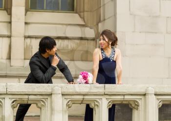 Horizontal Photo of young adult man looking at his lady friend, bouquet of flowers between them, with building in background 
