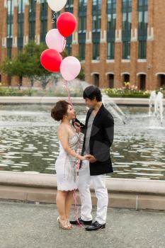 Vertical photo of young adult couple looking at each other, holding hands, with several balloons overhead of them with water fountain, flowers, trees and brick building in background 