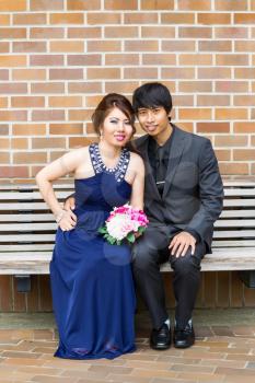 Vertical photo of young adult couple, looking forward, sitting on bench while holding each other with brick wall in background 