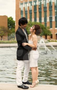 Vertical photo of young adult couple holding each other while standing on top of the water fountain ledge with trees, flowers and buildings in background 