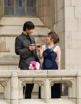 Vertical photo of young adult couple sharing a drink, bouquet of flowers between them, with building in background 
