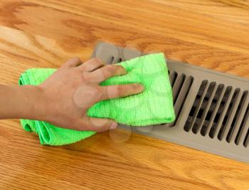 Horizontal photo of female hand cleaning outside grill plate of heater floor vent with Red Oak Floors in background 