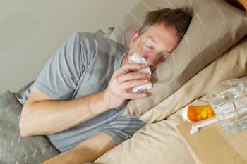 Closeup photo of mature man wiping nose with tissue while lying in bed with night stand and medicine in forefront 