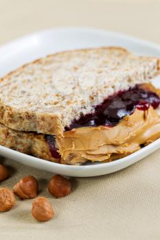 Closeup vertical photo of a peanut butter and jelly sandwich cut in half, inside white plate with whole nuts lying on textured table cloth 