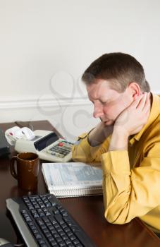 Vertical photo of mature man, holding head with hands, working on his taxes with tax booklet and office equipment in background 