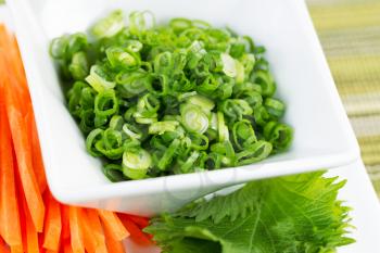 Closeup horizontal photo of fresh green onions, placed in white bowl, and sliced cuts of carrots used as sushi ingredients with textured table cloth underneath