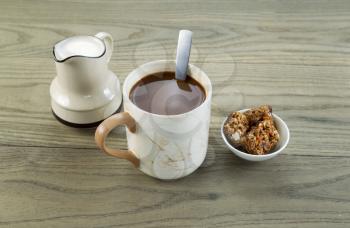 Closeup horizontal photo of a full cup of coffee, snacks and cream in pouring spout with aged wood underneath 