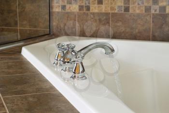 Horizontal photo of chrome faucet running water into soaking tub in master bathroom with partial shower glass in background 