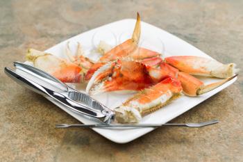 Horizontal photo of freshly cooked Dungeness crab legs, focus on large claw in center, on white plate with stainless crab crackers and stone counter top underneath  