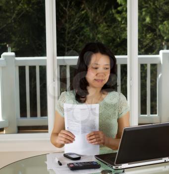 Photo of mature woman looking at computer screen, while holding tax forms, working at home with laptop, calculator and papers on top of table and large windows in background