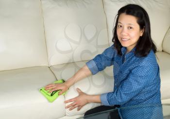 Horizontal photo of mature woman, looking forward, cleaning white leather sofa with microfiber rag in hand 