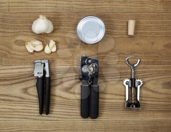 Overhead view of kitchen utilities placed on rustic wood.  Items including can opener, metal can, cork screw, cork, bottle opener, garlic press, and raw garlic. 