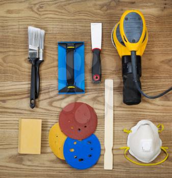 Overhead view of sanding and painting equipment positioned on rustic wooden boards.  Items include electric sander, mask, stir stick, sand paper, scrapper, sanding block, and brushes. 