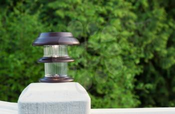 Horizontal photo of solar lamp, light just turning on, on patio post with green trees in background 