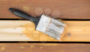 Horizontal photo of a large nylon paint brush on top of new wood board showing cedar stain color next to faded wood on outdoor deck 
