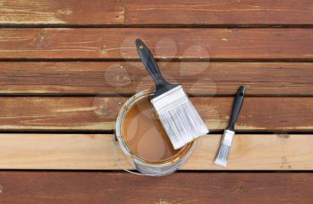 Horizontal photo of large nylon paint brush on top of wood stain can with small paint brush lying on a single new cedar wooden board next to fading wood on outdoor deck 