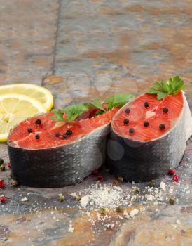 Closeup vertical photo of fresh red salmon steaks, course spices, herbs and lemon slices with natural stone slate underneath 