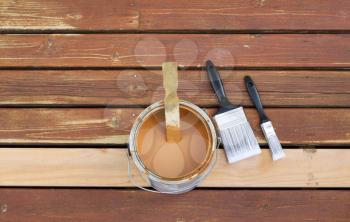 Horizontal photo of an open can of wood stain with two paint brushes lying on a single new cedar wooden board next to fading wood on outdoor deck 