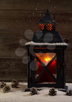 Vertical front view of an old lantern with glowing red candle inside surrounded by snow and rustic wood
