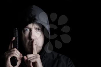 Closeup front view of mature man, looking forward and wearing hood, with weapon and index finger signaling to be silent on dark background  