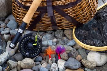 Top view up of  fishing fly reel, landing net, creel and assorted flies on dry river bed rocks 
