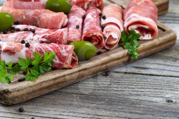 Close up horizontal image of various meats on serving board with ham, pork, beef, parsley, and olives on rustic wood. Focus on side part of serving board and first row of meat. 
