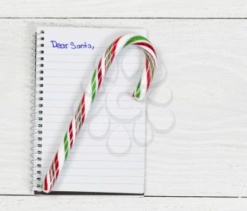 Candy cane on top of paper pad with writing to Santa on white desktop.
